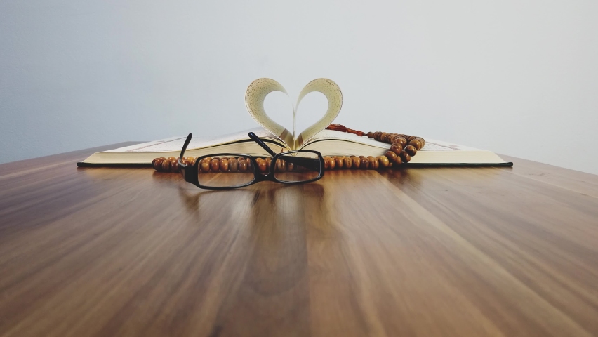 An approaching shot to an opened Quran with two sheets forming a heart shape, with reading glasses and the prayer beads in front of the Quran