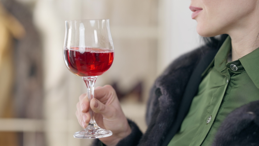 Red wine glass in female slim hand close-up. Unrecognizable Caucasian young woman shaking and tasting delicious luxurious drink indoors. Wealth and luxury concept