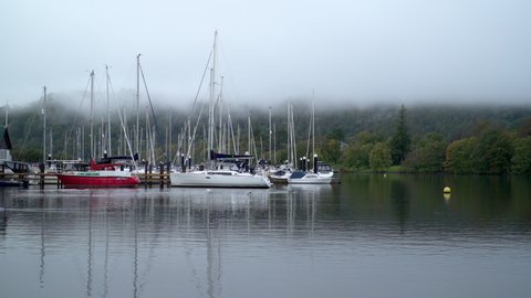 Magical marina with low cloud and mist, trees in the background and reflections in the water.  Sailing boats moored on Lake Windermere summer autumn 2020. Lake District, Cumbria, England. Tourism