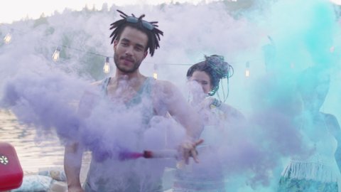 Joyous young male and female friends smiling at camera and dancing with colored smoke bombs during lake party on pier
