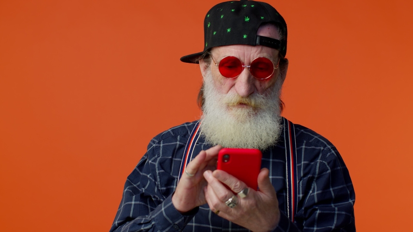 Happy excited joyful elderly bearded gray-haired man use mobile cell phone typing browsing say wow yes, found out great big win news doing winner gesture. Senior old grandfather on orange background Royalty-Free Stock Footage #1070630149