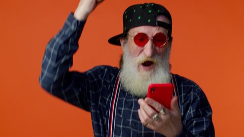 Happy excited joyful elderly bearded gray-haired man use mobile cell phone typing browsing say wow yes, found out great big win news doing winner gesture. Senior old grandfather on orange background