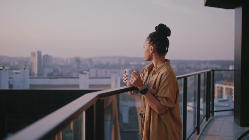 Young inspired woman standing on her terrace, enjoying drinking tea, watching views of sunrise landscape and scenery. People at home. Peaceful morning. | Shutterstock HD Video #1070643286
