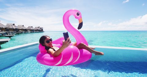 Phone - Travel vacation woman on inflatable Flamingo float mattress using app on mobile cell phone in swimming pool, maybe for booking hotel while relaxing sunbathing on resort pool in bikini