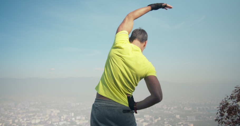 Older male is stretching and he exercises early in the morning on the mountain