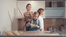 Blond children dancing, two girls and a boy having fun and dancing. Bright kitchen. On the table is a cookie dough.