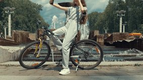 Beautiful fashionable girl makes a selfie on the phone while sitting on a bike. There is a large fountain in the background. Slow motion.