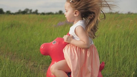 Little girl, daughter playing in field outdoors. Family and childhood. Happy little girl is playing in park and jumping on an inflatable toy donkey. Kid plays in field with his favorite toy horse.