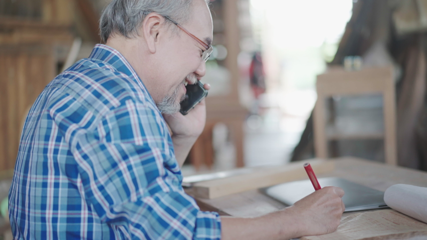 Asian senior male entrepreneur talking on mobile phone and listing order on paper at carpentry workshop studio.