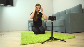 A beautiful athletic female fitness blogger talks to a mobile phone camera and records her Criscross Crunch workout in a spacious and bright living room with a cozy modern interior.