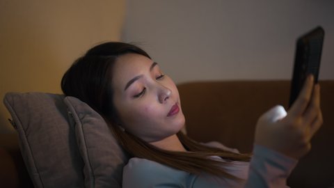 Young Asian woman laying down on the sofa feel boring with internet content or news in smartphone. Alone female stay at home have nothing to do at night. 