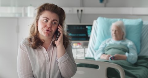 Young woman in hospital ward talking on smartphone with sick aged mother in hospital bed on background. Adult daughter having phone consultation with doctor visiting ill old mother in nursing home