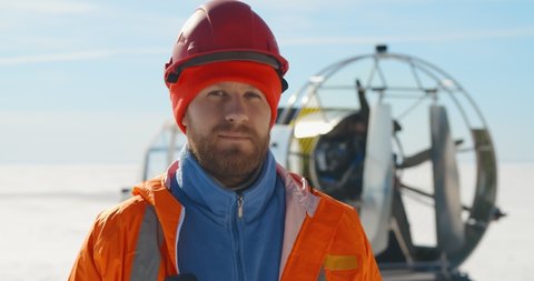 Close up portrait of handsome bearded lifeguard in uniform putting on hardhat posing at camera patrolling winter coast on hovercraft. Rescue service staff on frozen lake