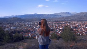 Young red hair woman traveler or hipster takes a video shot of the landscape in Montenegro. Mountains on the background. Sunny day. Hands on shot