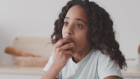 Healthy snack for kids. Close up portrait of thoughtful curly african american girl eating red apple, biting ripe fruit and smiling, sitting at home kitchen, slow motion