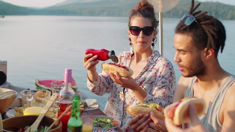 Company of cheerful multiethnic men and women eating hot dogs, chatting and smiling while having lake party on summer day