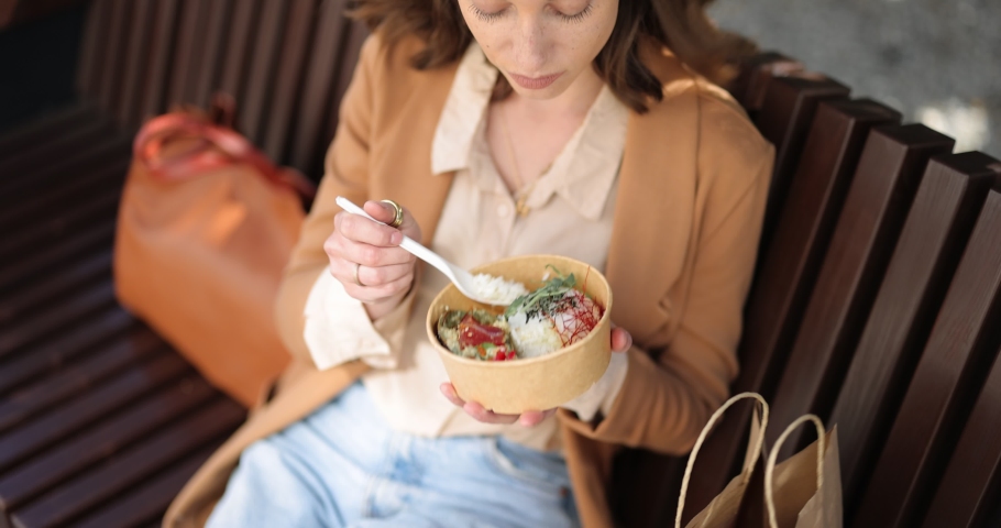Young businesswoman eating healthy takeaway food with tuna and rise, while sitting on the bench outdoors Royalty-Free Stock Footage #1070695948