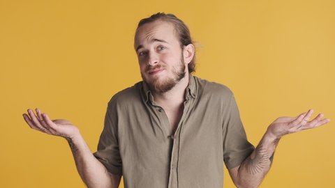 Young bearded man shrugging shoulders showing I do not know expression on camera over yellow background. I do not care