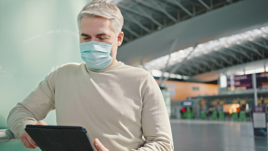 Travel portrait. Mature man wearing protective medical mask standing at airport hall with digital tablet, tracking shot, slow motion