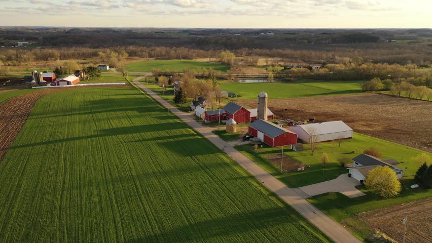 American midwestern countryside in springtime. Aerial view, drone flying over farms with red barns, rural road, agricultural fields. Sunny sky, clouds, spring