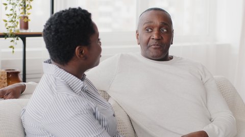 Family conversation at home, afro american couple husband and wife talking sitting on sofa, black ethnic mature middle aged man father listening nods head understand and adult african woman chatting