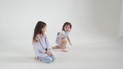 beautiful little girl with long hair in jeans and shirt posing against a white background and the younger sister runs by. children's photo shoot.