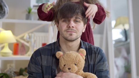 Portrait of funny young man looking at camera holding teddy bear as little girl making ponytail on father's hair. Joyful Caucasian father playing with daughter at home on weekend. Lifestyle and family