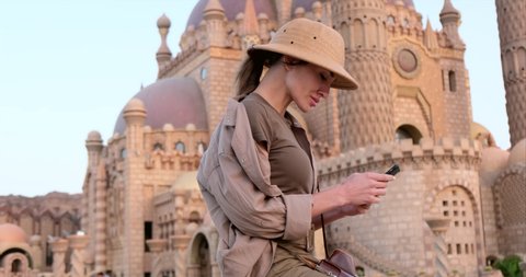 beautiful girl in travel hat, safari clothing writes a message on a mobile phone, India, against the background of the mosque .a traveler is resting after exploring the cultural attractions of india.