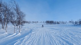 Close up video on two persons drive snowmobile through snowy landscape with birch trees in Lapland towards mountains. Cold sunny windy day