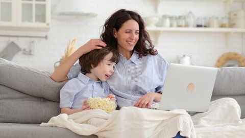 Smiling mother with little preschool son watch funny video, cartoon on laptop computer sit together on sofa in living room. Young mom and cute boy with popcorn under blanket spend time at funny movie