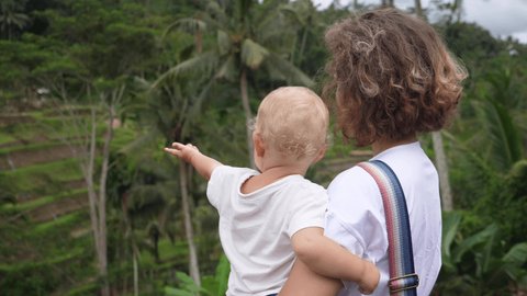 Back view of a Caucasian woman holding her baby looking at the jungle landscape.Blonde baby pointing far away 