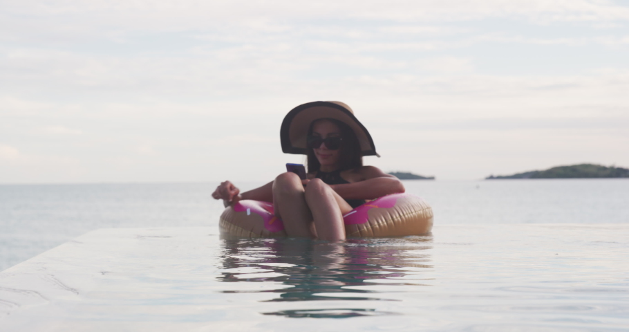 Vacation woman on inflatable donut float mattress using mobile cell phone in swimming pool. Girl relaxing relaxing enjoying travel holidays at resort infinity pool in bikini. Luxury lifestyle. Royalty-Free Stock Footage #1070722396