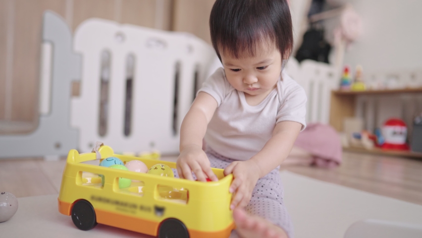 Asian cute baby girl playing on the wooden floor in the playroom, with her mother closely supervised during the day, cute playful daughter trying to reach balls to fill on the yellow car toy in hand