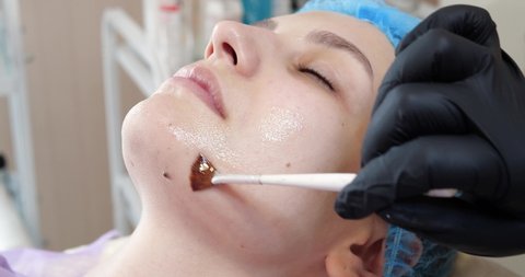 Cosmetologist is applying peeling mask on woman's face using brush in beauty clinic. Beautician making skincare procedure. Spa treatment in cosmetology