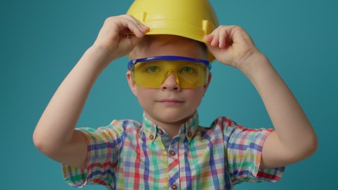 Cute preschool boy wearing protective eyeglasses putting on yellow helmet looking at camera. Young kid builder on blue background. 