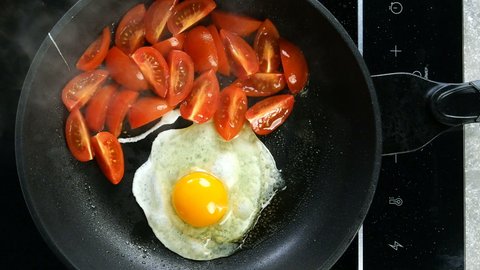 Preparation fried eggs with tomato in frying pan for breakfast on electric stove on domestic kitchen. Raw broken chicken egg falls into hot skillet. Top view. Top down food. Close-up.