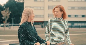 Females colleagues who are talking, communicating, town street. Woman riding scooter. Outdoor business meeting. Blonde and Redhead woman friends walking on autumn day. 4K video.
