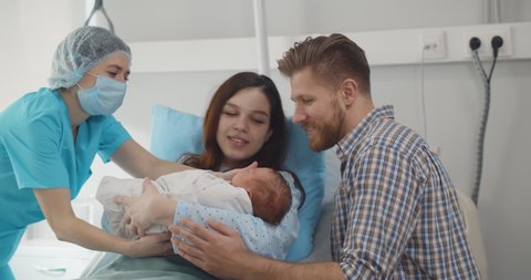 Young caucasian couple with doctor holding newborn baby in hospital ward. Nurse wearing protective mask giving infant to happy parents in clinic room. Healthcare concept