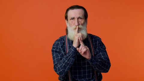 Shh be quiet please! Elderly bearded gray-haired man presses index finger to lips makes silence gesture sign do not tells secret. Senior old grandfather on orange background. People lifestyle emotions