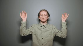 Nerd in glasses and shirt, standing on a gray wall background and meditating with his eyes closed and calm face. Young male student engaged in meditation, yoga concept