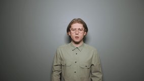 Shocked young man in glasses and shirt stands on a background of gray gray wall and points his finger to the side on copy space