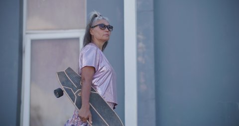 Cheerful healthy old senior woman holding skateboard and walking down street with happy emotion. Cool elderly asian granny enjoy summer outdoor activity play extreme sport surf skate in slow motion.