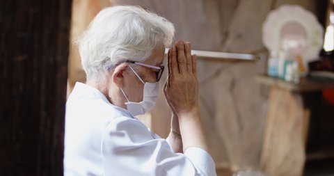 An Asian old lady, a Buddhist, has white hair, wearing glasses and a mask to prevent COVID-19 , sitting and hands clasping, listen to Buddhist monks praying at the merit making ceremony in the temple.