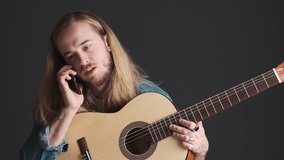Long haired blond guy with acoustic guitar talking with friend on smartphone after rehearsal in studio isolated on black background