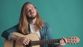 Young long haired blond bearded man playing on acoustic guitar and singing new song looking inspiring over colorful background. Music concept