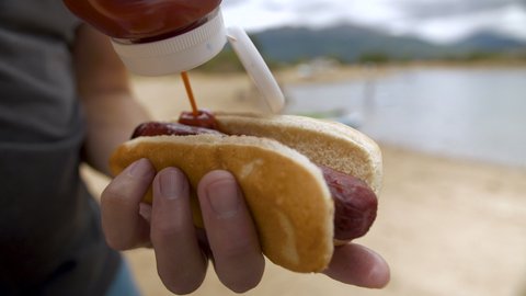 putting ketchup on a hot dog or bratwurst during a picnic on the beach by a lake with mountain views.