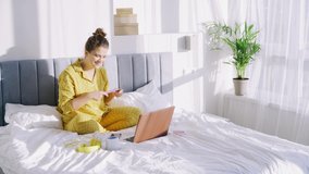 Attractive young woman in yellow pajamas with breakfast put on bed applies cream on face watching video on laptop at home in bright sunny morning