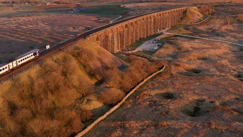 Aerial drone footage of moving train over long viaduct with arches, taken at sunrise. Ribblehead Viaduct, Yorkshire Dales, UK.