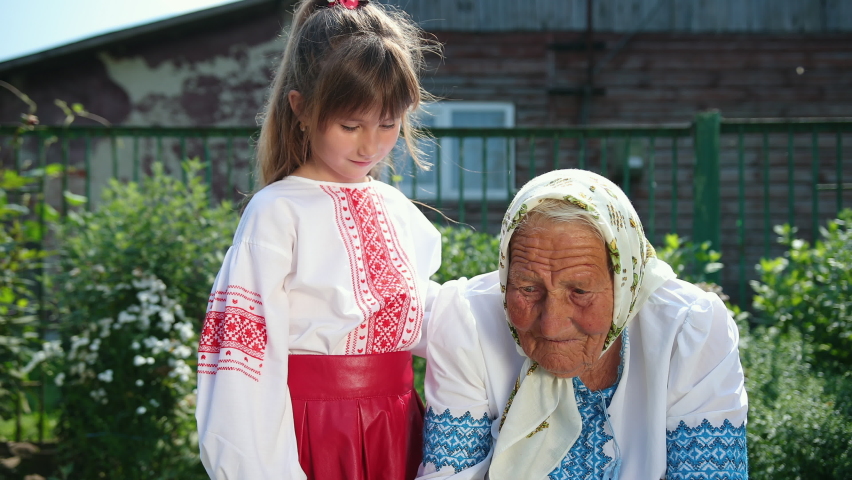 An old grandmother is sitting on the street and reading a book. She is dressed in national ukrainian clothing vyshyvanka. Girl listen carefully to their grandmothers who read fairy tales