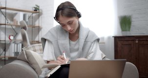 Focused Girl Learning Distance listening Lecture in Headset and writing Notes. Attractive Student having Online Webinar, Training looking at Laptop screen while sitting at Home. Quarantine Concept.